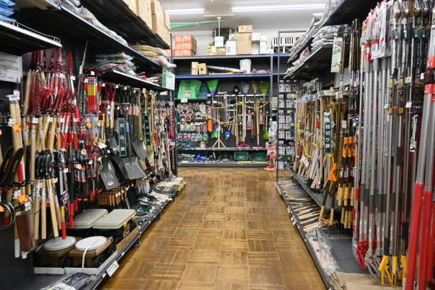 Allée de quincaillerie bordée de manches d’outils de jardin et de balais, plancher de bois au centre